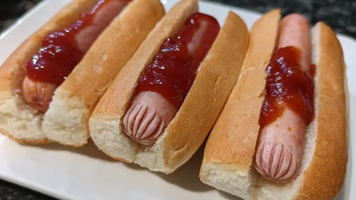 three hot dogs in buns topped with ketchup on a white plate